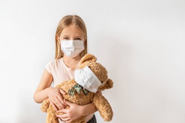 Coronavirus ve Hava kirliliği pm2.5 konsepti. Karantina sırasında oyuncak ayısını korumak ve tutmak için maske takan küçük Rus kız. Önleme salgını. Yurt içi kampanyada kal .