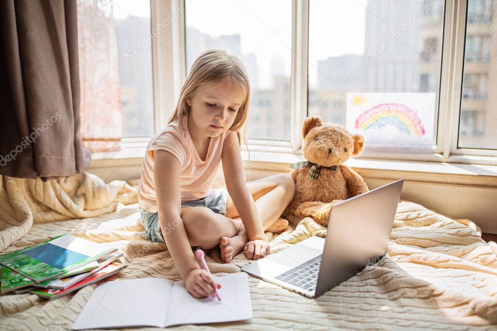 Pretty schoolgirl studying homework during her online lesson at home, social distance during quarantine covid-19, self-isolation, online education concept, home schooler. Workplace in living room