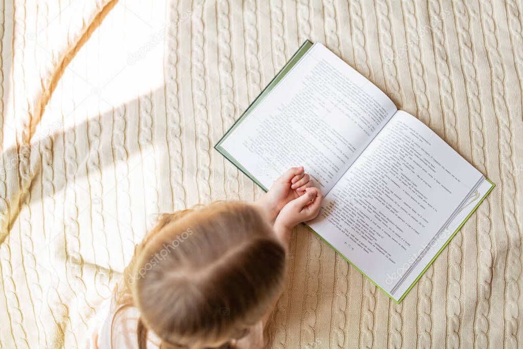 Cute little caucasian girl in casual clothes reading a book. Kid lying on a bed in the room. Stay at home during coronavirus covid-19 pandemic, distance education time. Overhead, top view, flat lay.