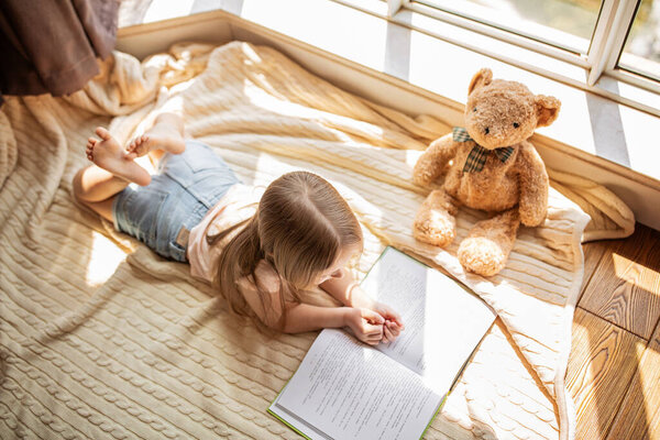Cute little caucasian girl in casual clothes reading a book with stuffed teddy bear toy and smiling while lying on a floor near window in the room. Stay at home during coronavirus covid-19 pandemic.