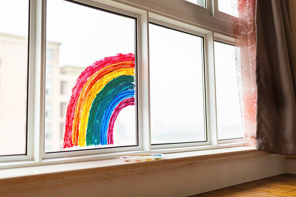 Kid painted rainbow during Covid-19 quarantine at home. Girl near window. Stay at home Social media campaign for coronavirus prevention, let's all be well, hope during coronavirus pandemic concept.