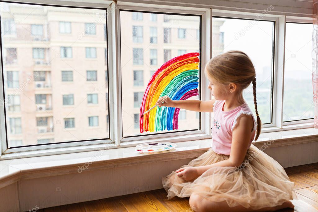 Kid painting rainbow during Covid-19 quarantine at home. Girl near window. Stay at home Social media campaign for coronavirus prevention, let's all be well, hope during coronavirus pandemic concept.