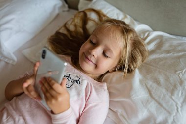 Akıllı telefondan videolar izleyen sevimli küçük bir kız. Yatakta tek başına gülen yüzlü bir çocuk. Evde mutlu yüzlü cep telefonu kullanan bir çocuk. Evde karantina Coronavirus COVID-19 pandemik önleme