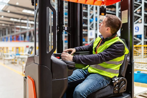 Storehouse man employee in uniform using forklift with box in modern automatic warehouse. Boxes are on the shelves of the warehouse. Warehousing, machinery concept. Logistics in stock.