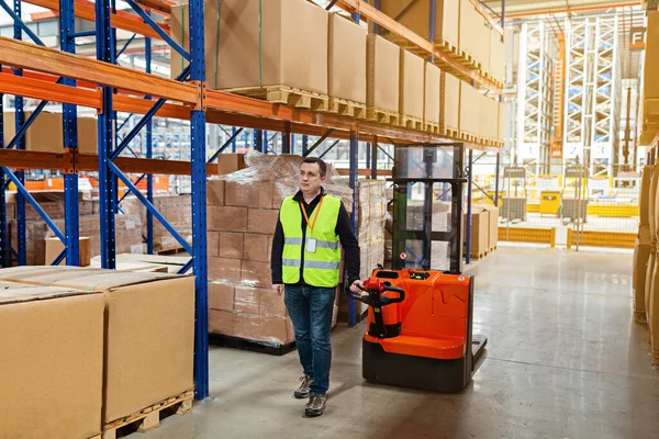 Storehouse man employee in uniform using forklift with box in modern automatic warehouse. Boxes are on the shelves of the warehouse. Warehousing, machinery concept. Logistics in stock.