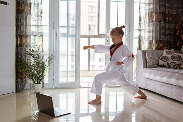 Taekwondo chica en kimono con cinturón blanco haciendo ejercicio en casa en la sala de estar. Educación en línea durante el bloqueo del coronavirus covid-19, autoaislamiento y concepto de distanciamiento social — Foto de Stock