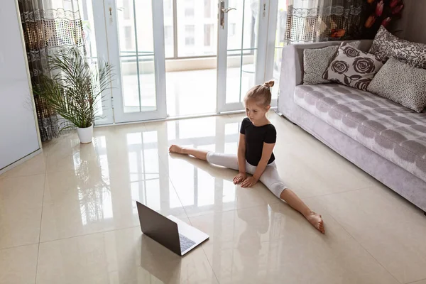 Cute little girl in sportswear watching online video on laptop and doing fitness exercises at home. Distant training with personal trainer, social distance or self-isolation, online education concept.