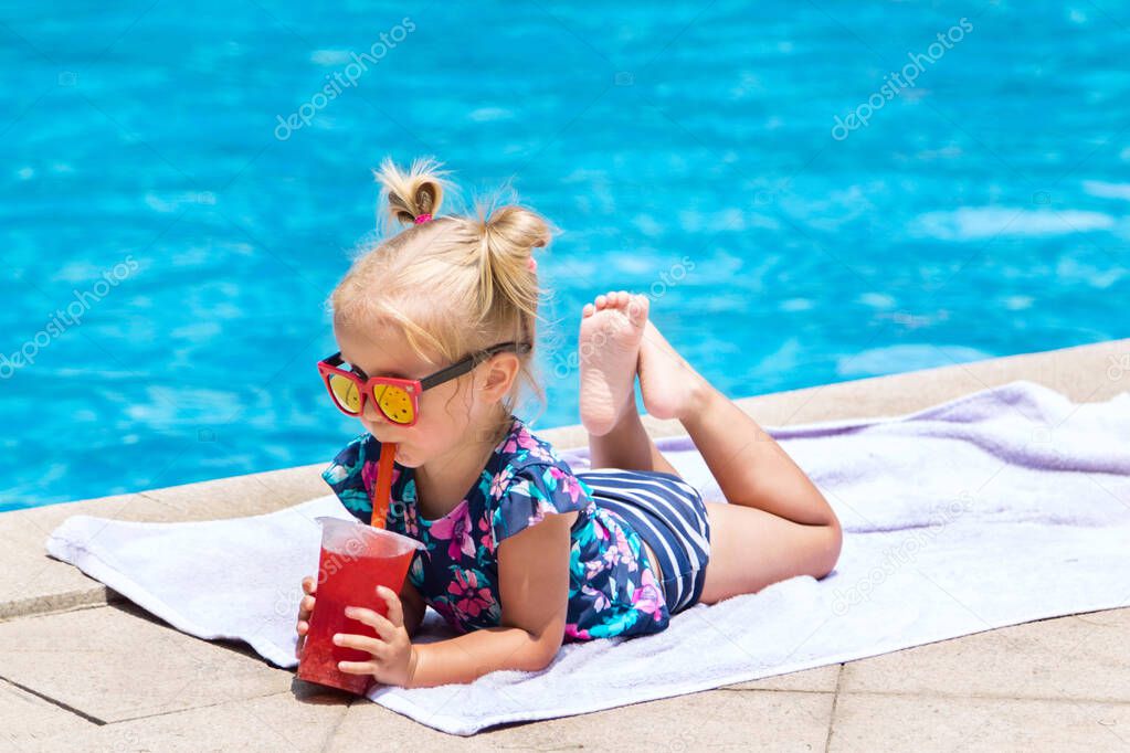Beautiful little girl, cute toddler with blonde hair in swimwear, sitting at a swimming pool and drinking water melon juice with fresh fruit. Kid having fun during vacation in a tropical resort.