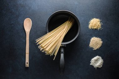 Makarnalı küçük bir tencere, ahşap bir yemek kaşığı ve kırsal bir masada çeşitli çiğ makarna yığınları. Geleneksel İtalyan mutfağı kavramı. Moody tarzı