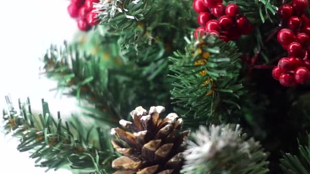 Καλά Χριστούγεννα και καλή χρονιά! Πράσινο χριστουγεννιάτικο δέντρο ελαφρά ξεσκονισμένο με χιόνι. Πλαστικό δέντρο σε ελαφρύ φόντο με τεχνητά μούρα και φυσικά κουκουνάρια "επιπλέει" μπροστά από το φακό. — Αρχείο Βίντεο