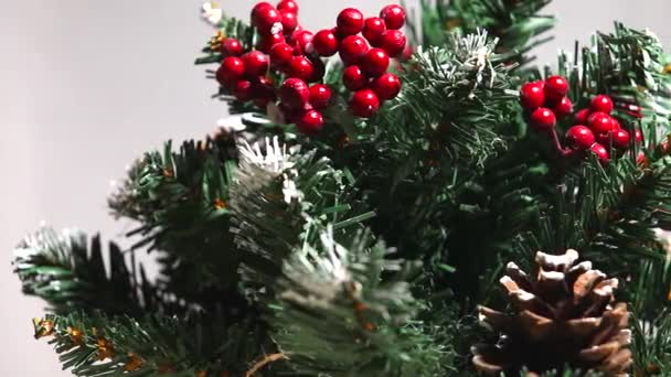 Feliz Navidad y un feliz año nuevo! Árbol de Navidad verde ligeramente espolvoreado con nieve. Árbol de plástico sobre un fondo claro con bayas artificiales y conos de pino natural "flota" delante de la lente . — Vídeo de stock