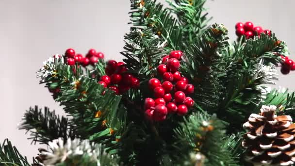 Merry christmas and a happy new year! Green Christmas tree lightly dusted with snow. Plastic tree on a light background with artificial berries and natural pine cones "floats" in front of the lens. — ストック動画