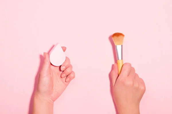 Woman holding makeup brush and beauty blender on pastel pink background. Beauty accessories with makeup brush versus beauty sponge for foundation. Beauty flat lay, top view, copy space
