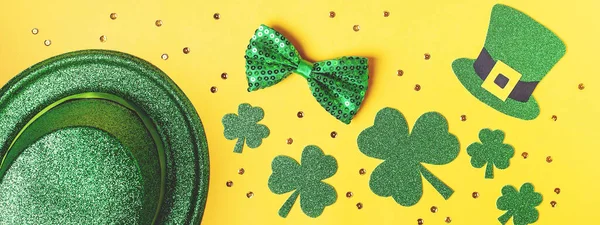 Saint Patrick's day holiday card with green shamrock symbols, hat, golden confetti. Traditional St. Patrick's Day green attire and decorations on yellow background. Web banner, copy space, top view