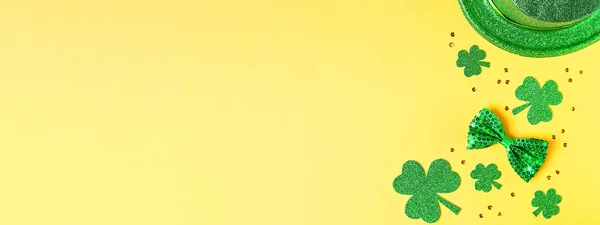 Saint Patrick\'s day holiday card with green shamrock symbols, hat, golden confetti. Traditional St. Patrick\'s Day green attire and decorations on yellow background. Web banner, copy space, top view