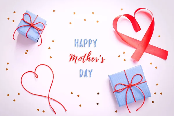 Happy Mother\'s Day greeting card with red hearts, blue gift boxes and golden confetti on pastel table top. Background for Mothers Day holiday with text
