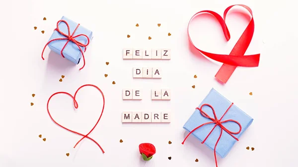 Happy Mother\'s Day greeting card with red hearts, blue gift boxes and golden confetti on pastel table top, overhead shot. Feliz dia de la madre text from wooden blocks that mean Happy Mothers day