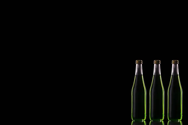 Three small glass bottles with a green drink stand on a mirror surface against a dark background with the right.