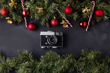 Kırmızı toplarla süslenmiş kozalaklı Noel çelengi ve siyah masanın üst ve alt kısmında karamel kamışlarla süslenmiş altın çanlar ve ortada da klasik bir kamera..