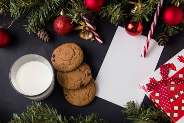Koyu bir masa üzerinde koni ve süslemeli Noel ağacı dalları hediye tepsisi ve kurabiyeli sütün yanında..