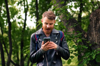 Kızıl saçlı sakallı bir adam ormanın ortasında dururken akıllı telefon ekranına bakarken geniş bir gülümseme sergiliyor..