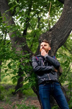 Siyah deri ceketli ve mavi kotlu bir adam düşünceli bir şekilde uzaklaşıyor, sırtını ağaca dayıyor ve elleriyle sakalını sallıyor..