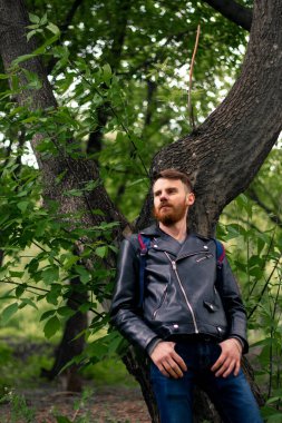 Siyah deri ceketli bir adam, arkasını ağaca dayamış, düşünceli bir şekilde uzaklara bakar..