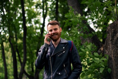 Kırmızı sakallı bir adam, siyah deri ceketli yeşil bir ormanda dururken akıllı bir telefonla konuşurken geniş bir gülümseme sergiliyor..