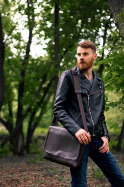 Omzunda kahverengi bir çanta ve kalın sakallı genç bir İrlandalı ormanın ortasında dikilirken mesafeye bakar..