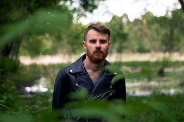 Turuncu sakallı ve bıyıklı şık bir adam yeşil bir ormanda çim ve dallardan oluşan bir arka planda duruyor..