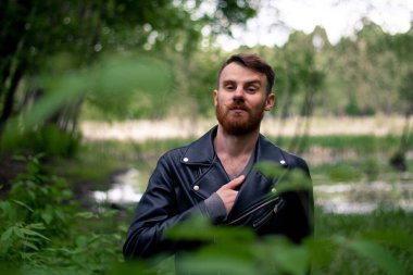 Sakallı ve bıyıklı şık bir adam yeşil bir ormanda duruyor. Arka planda çimenler ve deri ceketli dallar var. Eli göğsünde..