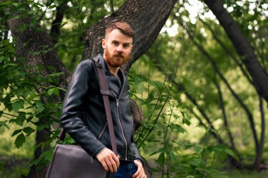 Eli çantasında olan adam parkta büyük bir ağacın yanında duruyor..