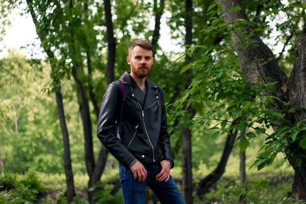 A guy with a red mustache in a leather jacket and blue jeans stands in the forest with his hands in the feed.