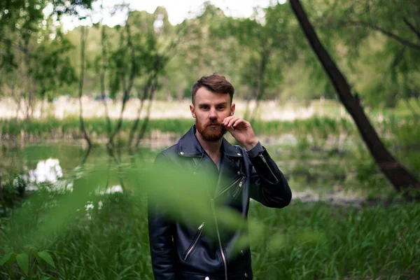 A young guy with a red mustache and a beard trembles with his mustache against a green forest looking to the right with foliage in the foreground.