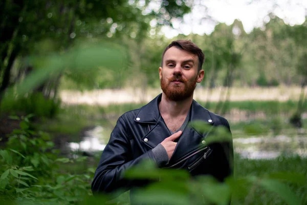 Sakallı ve bıyıklı şık bir adam yeşil bir ormanda duruyor. Arka planda çimenler ve deri ceketli dallar var. Eli göğsünde..