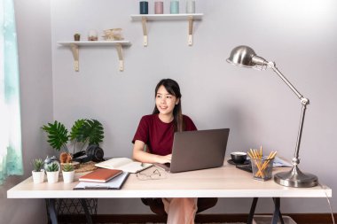 Genç, güzel, rahatlatıcı Asyalı bir kız evden uzakta masa başında çalışan bir dizüstü bilgisayar kullanıyor, Coronavirus için karantina ve akıllı çalışma konsepti.