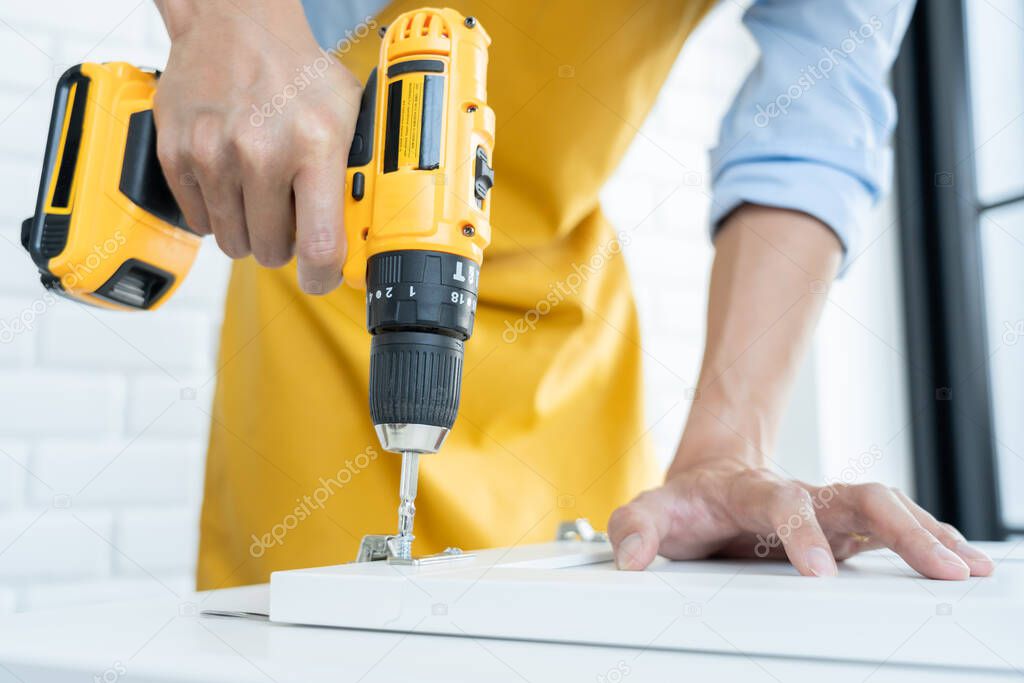 Close up portrait and details of caucasian male worker using electric screwdriver instrument in hand and repairing new wooden desk, home improvement concept