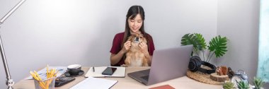 Evinde dizüstü bilgisayarla çalışan Asyalı güzel bir kadın. Oturma odasındaki koltukta ya da kanepede çalışıyor. Evcil köpek yavrusu gibi şirin bir köpek ve gardiyanla.