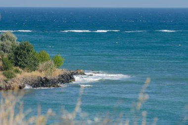 Uçurumdan büyüyen çimlerin arasından deniz kıyısına. Mavi okyanusta körfez, gün ortasında küçük beyaz dalgalar. Suyun ortasında yeşil çalıları olan taştan bir sahil. Deniz konsepti.