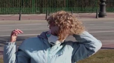 Genç kıvırcık sarışın saçını düzeltiyor ve sokakta tıbbi koruyucu yüz maskesi takıyor.