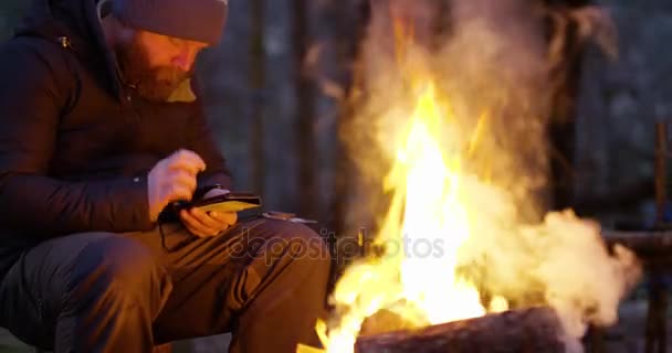 Ein Mann mit Kompass und Smartphone am Lagerfeuer im Wald — Stockvideo