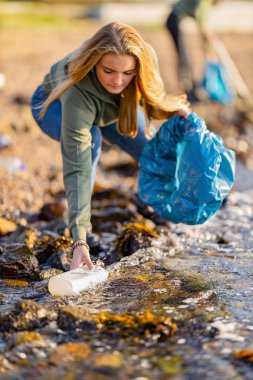 Young woman picking up garbage from rocky shore clipart