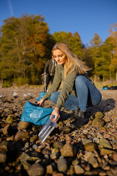 Young woman picking up plastic at rocky beach — Stok fotoğraf