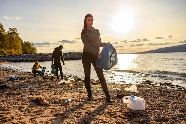 Mujer limpiando la playa con voluntarios durante la puesta del sol — Foto de Stock