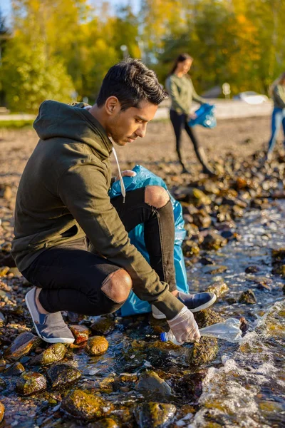 Young volunteer man crouching while picking up garbage on shore — Stok fotoğraf