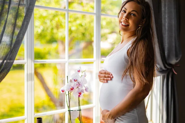 Sorridente donna incinta in piedi vicino alla finestra e toccando la pancia — Foto Stock