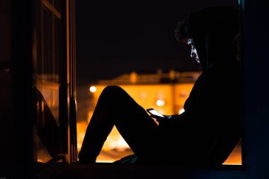 Cep telefonu olan genç bir adam gece pencereden dışarı bakıyor. Koronavirüs enfeksiyonunu önlemek için karantinaya alınmış..