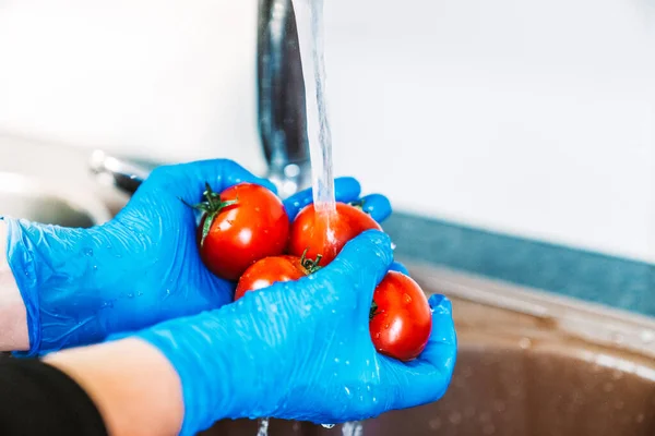 Hands with blue latex gloves disinfecting tomatoes to decontaminate the fruit from coronavirus. Washing the fruit with water in the kitchen sink with the tap.