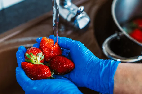 Hands with blue latex gloves disinfecting strawberries to decontaminate the fruit from coronavirus. Washing the fruit with water and lye in the kitchen sink.