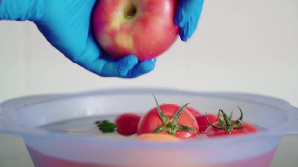 台所で水と漂白剤と青いプラスチック製のボウルに赤いリンゴを洗う青いラテックス手袋を手 花冠ウイルスの拡散を防ぐために果物を消毒する — ストック動画